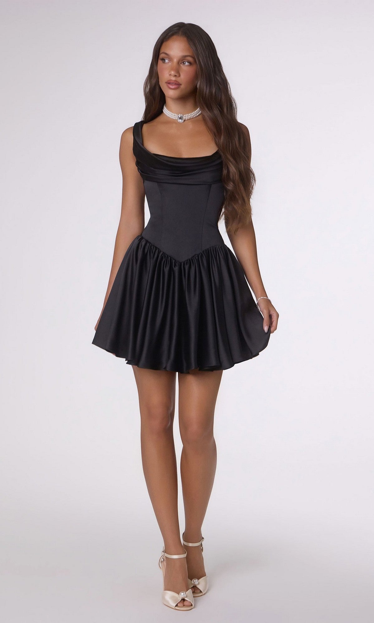Woman in the Amoris T735 short satin black homecoming dress with a corset bodice and A-line skirt, paired with white heels, a silver necklace, and long wavy hair, standing against a plain light background.
